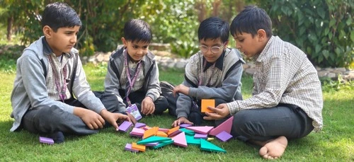 Four school boys in uniform sitting on grass outdoors, collaborating and playing with colorful geometric learning blocks.