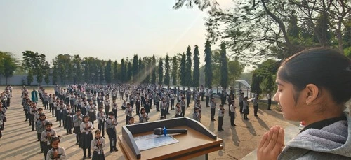 School students standing in rows during morning assembly while a girl leads prayer from the podium outdoors.