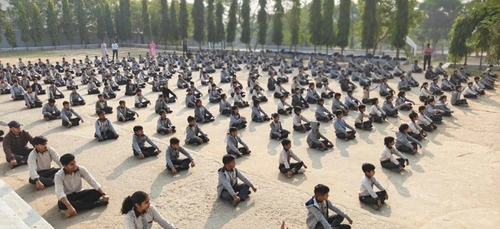 School students sitting cross-legged in organized rows during outdoor morning assembly in the school courtyard.